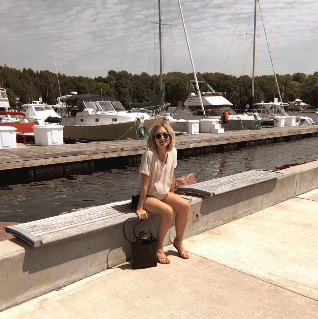 boats in the marina at egg harbor in door county, girl sitting by the dock with sailboats behind her, maternity t-shirt in a neutral color, crocodile bucket bag, blonde shoulder length straight bob